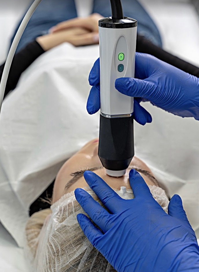 Zabieg na twarz z użyciem urządzenia kosmetycznego; widoczne dłonie w niebieskich rękawiczkach oraz twarz kobiety w czepku ochronnym. Urządzenie ma włączone zielone światło.