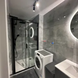 Nowoczesna łazienka z szarą, kwadratową glazurą, czarną kabiną prysznicową i pralką. Oświetlenie punktowe i okrągłe lustro z podświetleniem LED.