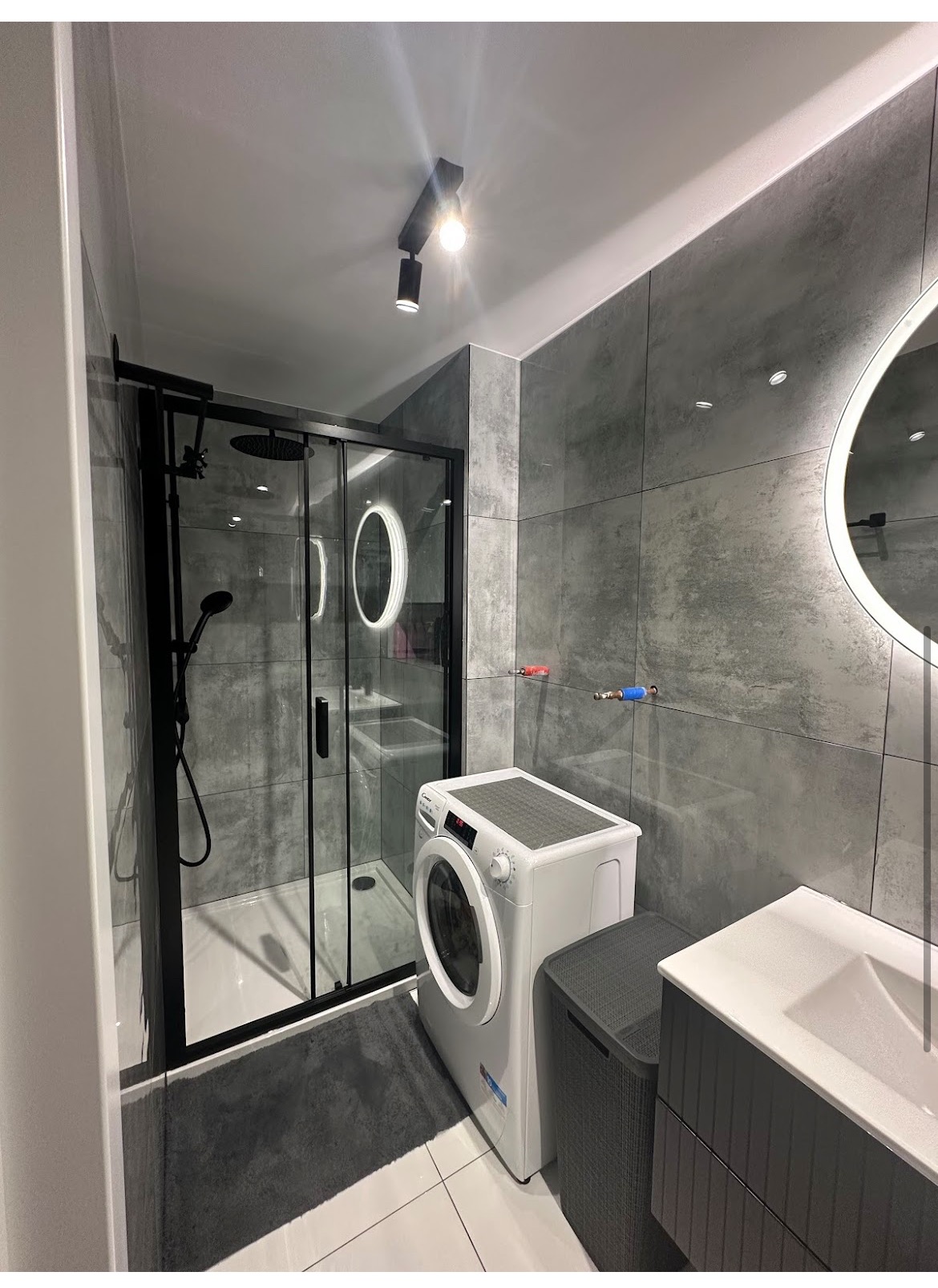 Nowoczesna łazienka z szarą, kwadratową glazurą, czarną kabiną prysznicową i pralką. Oświetlenie punktowe i okrągłe lustro z podświetleniem LED.