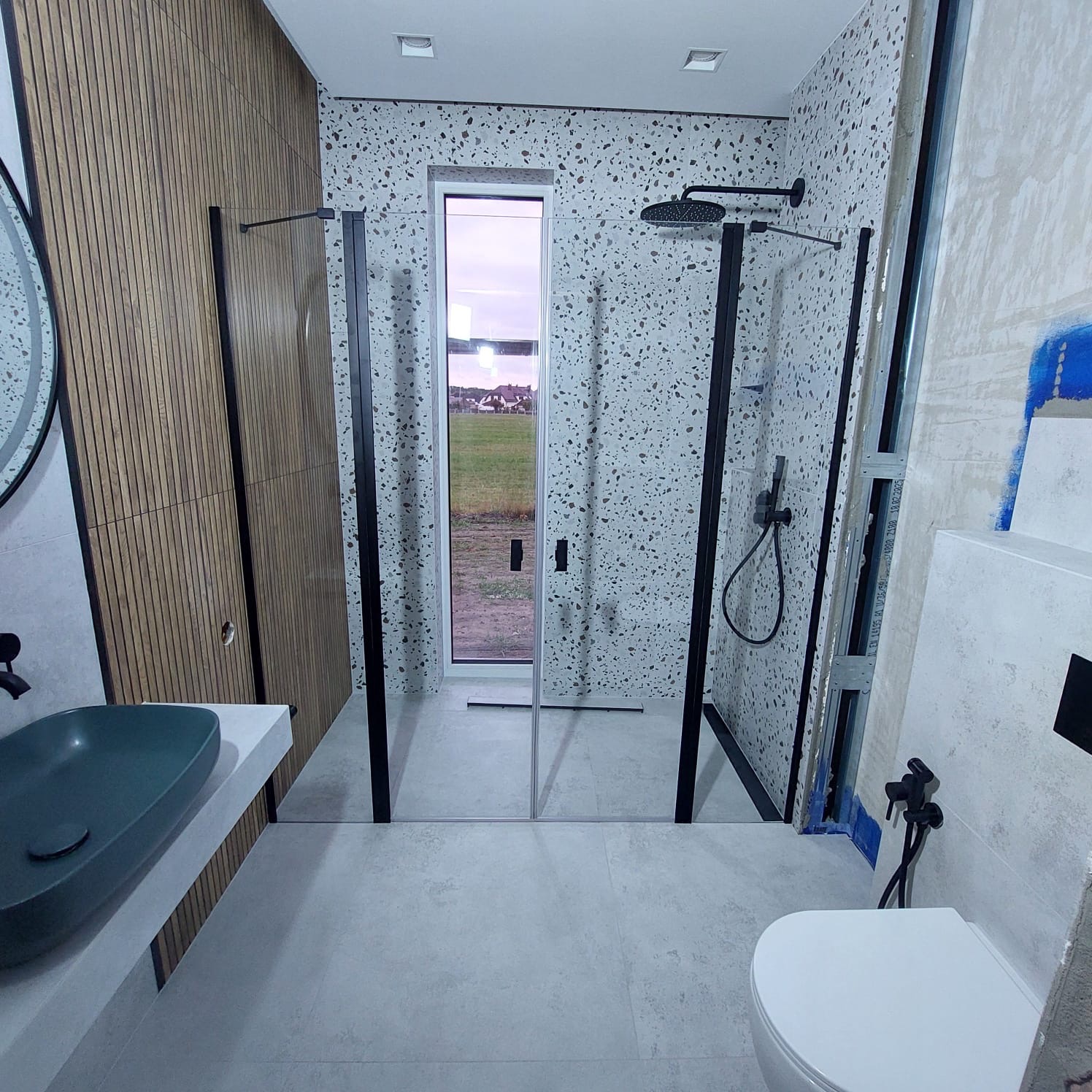 Nowoczesna łazienka z kabiną typu walk-in, umywalką w kolorze butelkowej zieleni i drewnianymi panelami ściennymi. Widok na zewnątrz przez okno w kabinie.