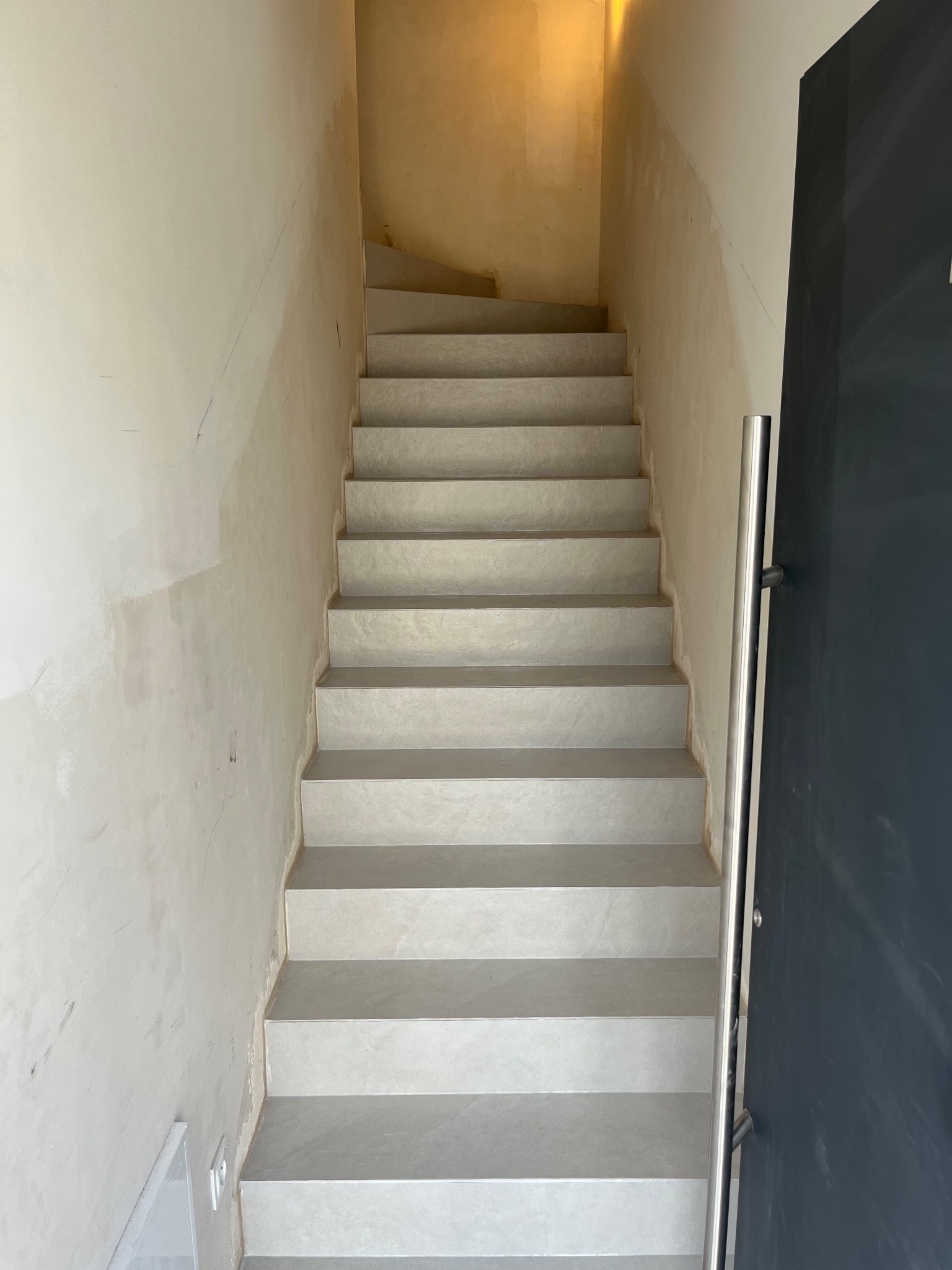Wykończone schody z szarymi płytkami, widoczne ślady po remoncie na ścianach i fragment ciemnych drzwi z metalowym uchwytem. Ujęcie z dołu schodów.