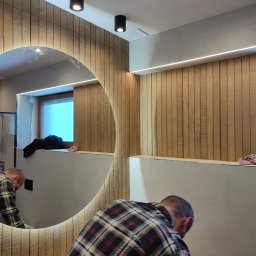Piotr Cierach - Wykończona łazienka z okrągłym lustrem podświetlonym LED, drewnianą okładziną ścienną i czarnymi lampami sufitowymi. Osoba w tle wykonuje prace wykończeniowe.