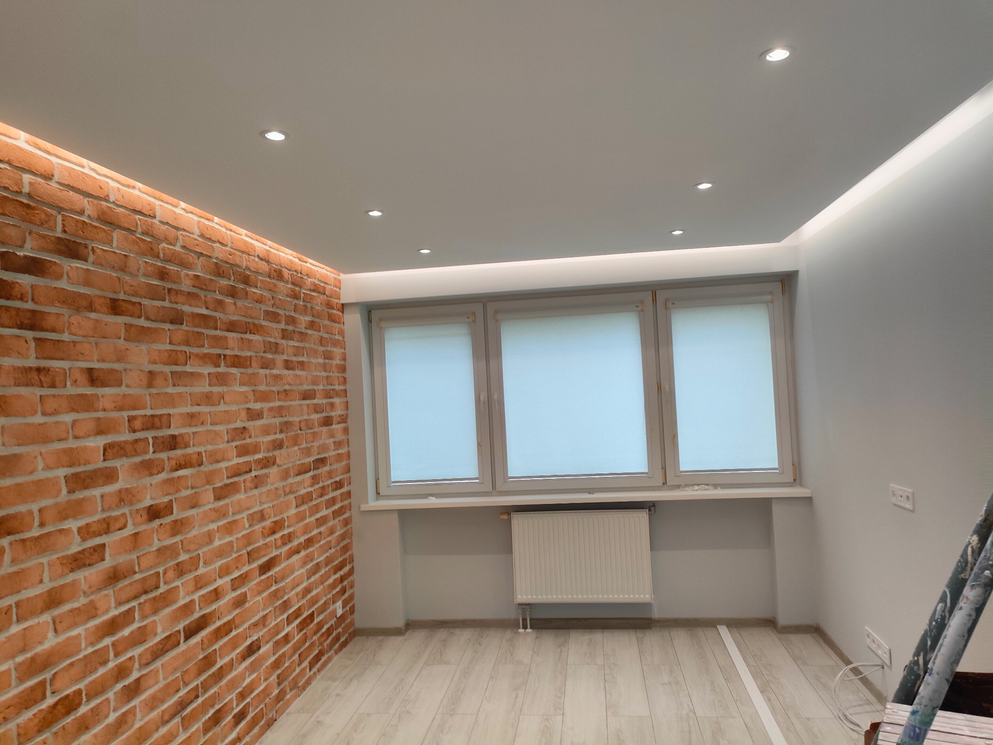 Pokój z ceglaną ścianą, trzema oknami z roletami i grzejnikiem. Sufit z oświetleniem punktowym i listwą LED. Podłoga laminowana. Widoczne ślady po malowaniu.