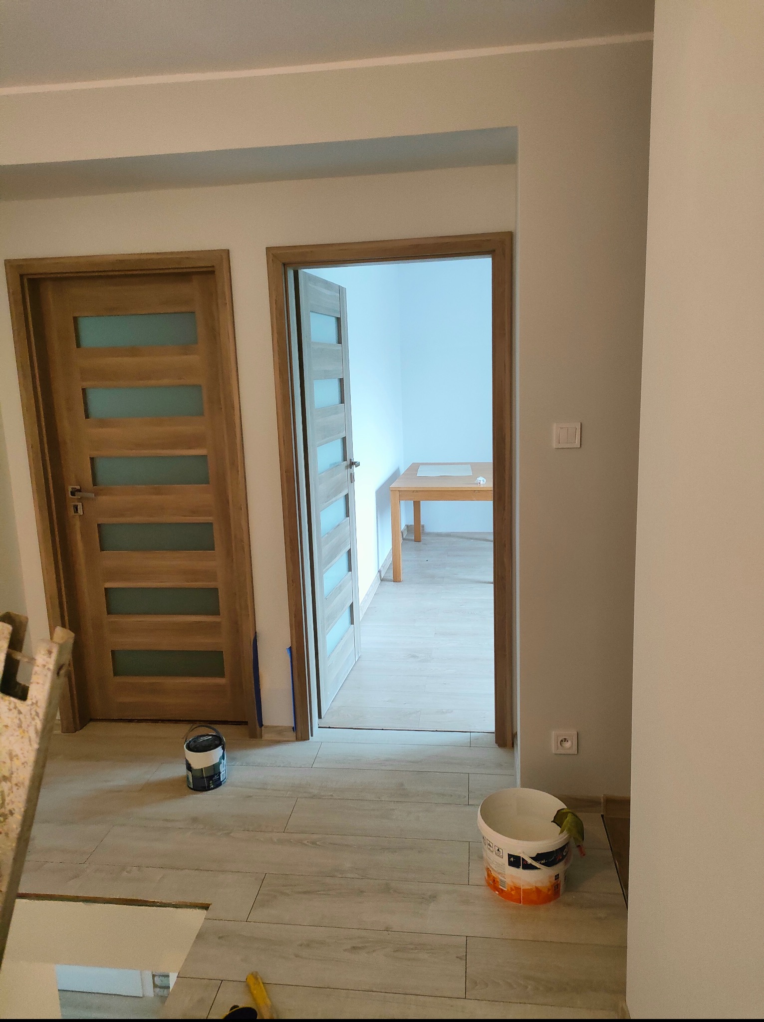 Jasne wnętrze po remoncie: drewniane drzwi z przeszkleniami, pomalowane ściany w odcieniach szarości, ceramiczna podłoga imitująca drewno, puszki z farbą.