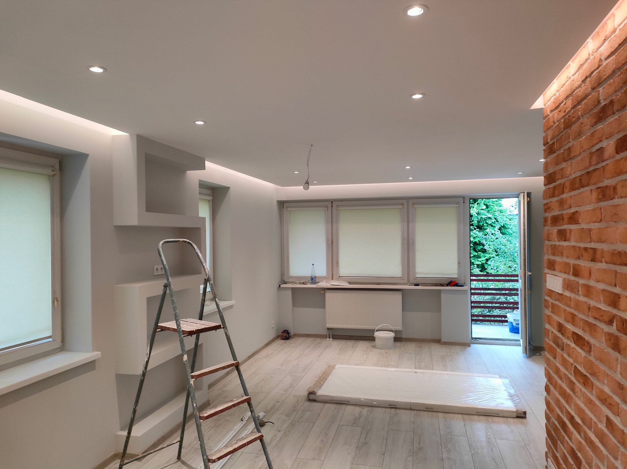 Jasne wnętrze po remoncie z ceglaną ścianą, drabina malarska i przygotowane podłoże. Widoczne oświetlenie sufitowe i szare ściany. Parkiet na podłodze.