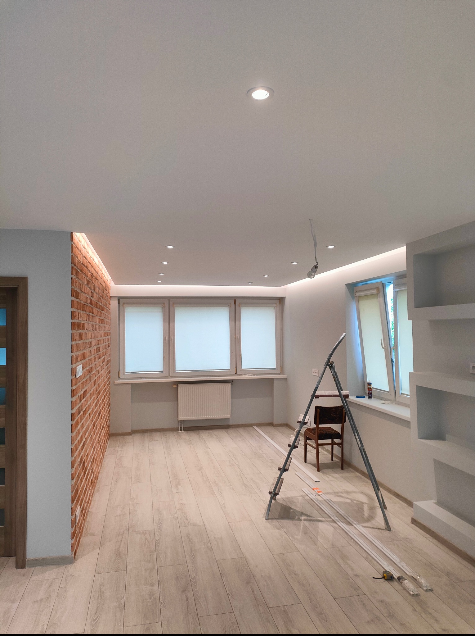 Jasne wnętrze po remoncie: ściana z cegły, drewniana podłoga, białe ściany i sufit z oświetleniem LED. Drabina i listwy przypodłogowe leżą na podłodze.