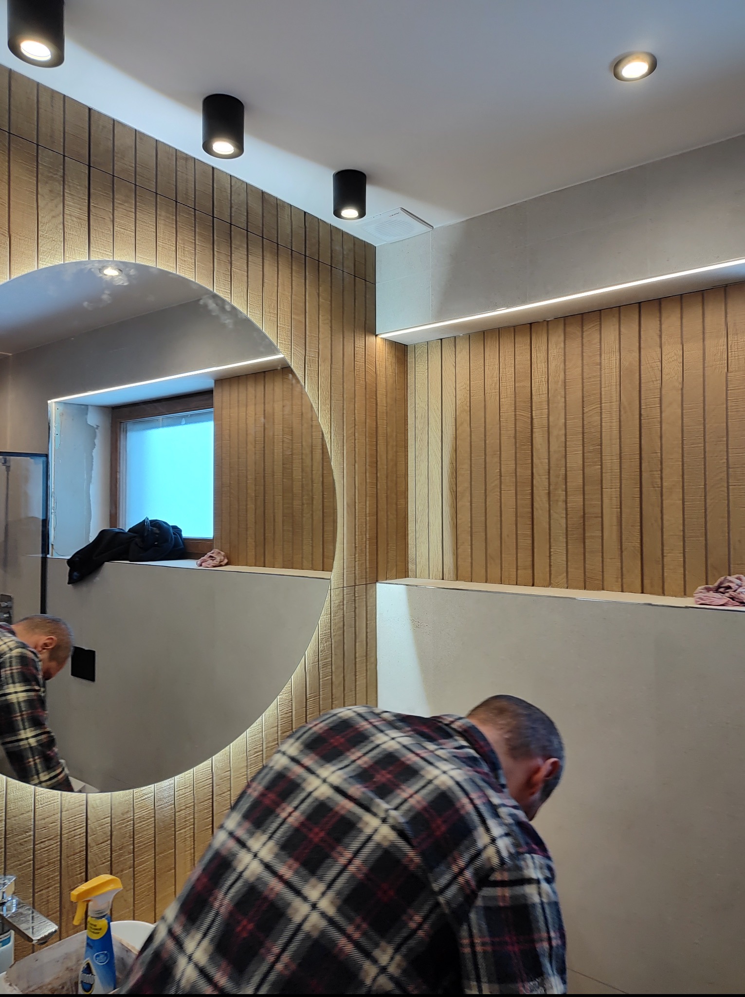 Wykończona łazienka z okrągłym lustrem podświetlonym LED, drewnianą okładziną ścienną i czarnymi lampami sufitowymi. Osoba w tle wykonuje prace wykończeniowe.