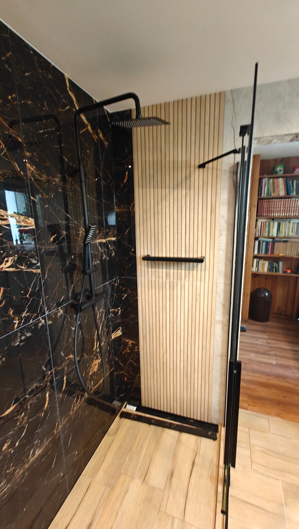 Nowoczesna kabina prysznicowa z czarną armaturą i panelami ściennymi imitującymi drewno. Widoczna marmurowa ściana i fragment półki z książkami w tle.