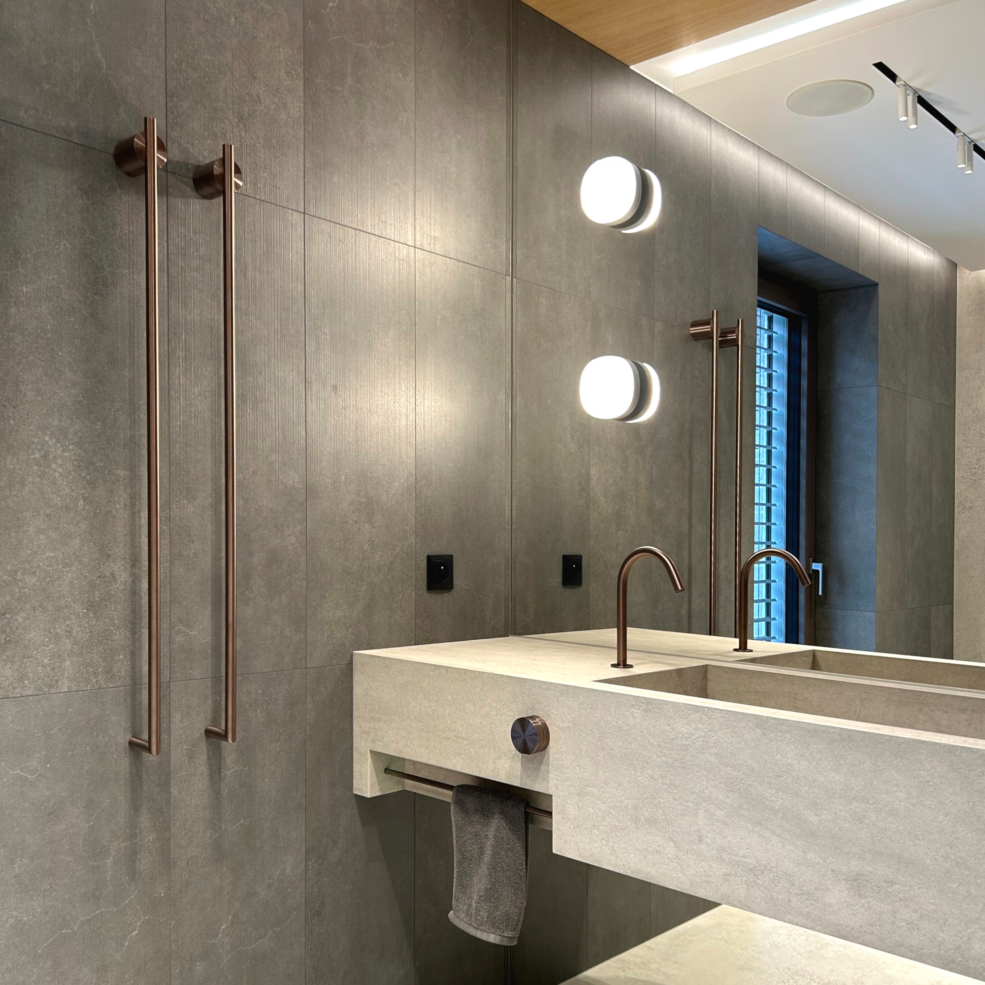 Nowoczesna łazienka z szarą płytką, podwójną umywalką z betonu architektonicznego i miedzianymi akcentami: baterie, grzejnik drabinkowy i oświetlenie.