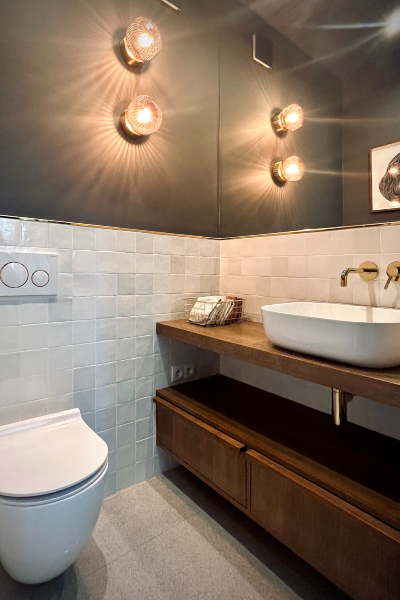 Nowoczesna łazienka z białymi płytkami, drewnianą szafką pod umywalką i złotymi akcentami, oświetlona przez dwa kinkiety z żarówkami o ciepłej barwie.