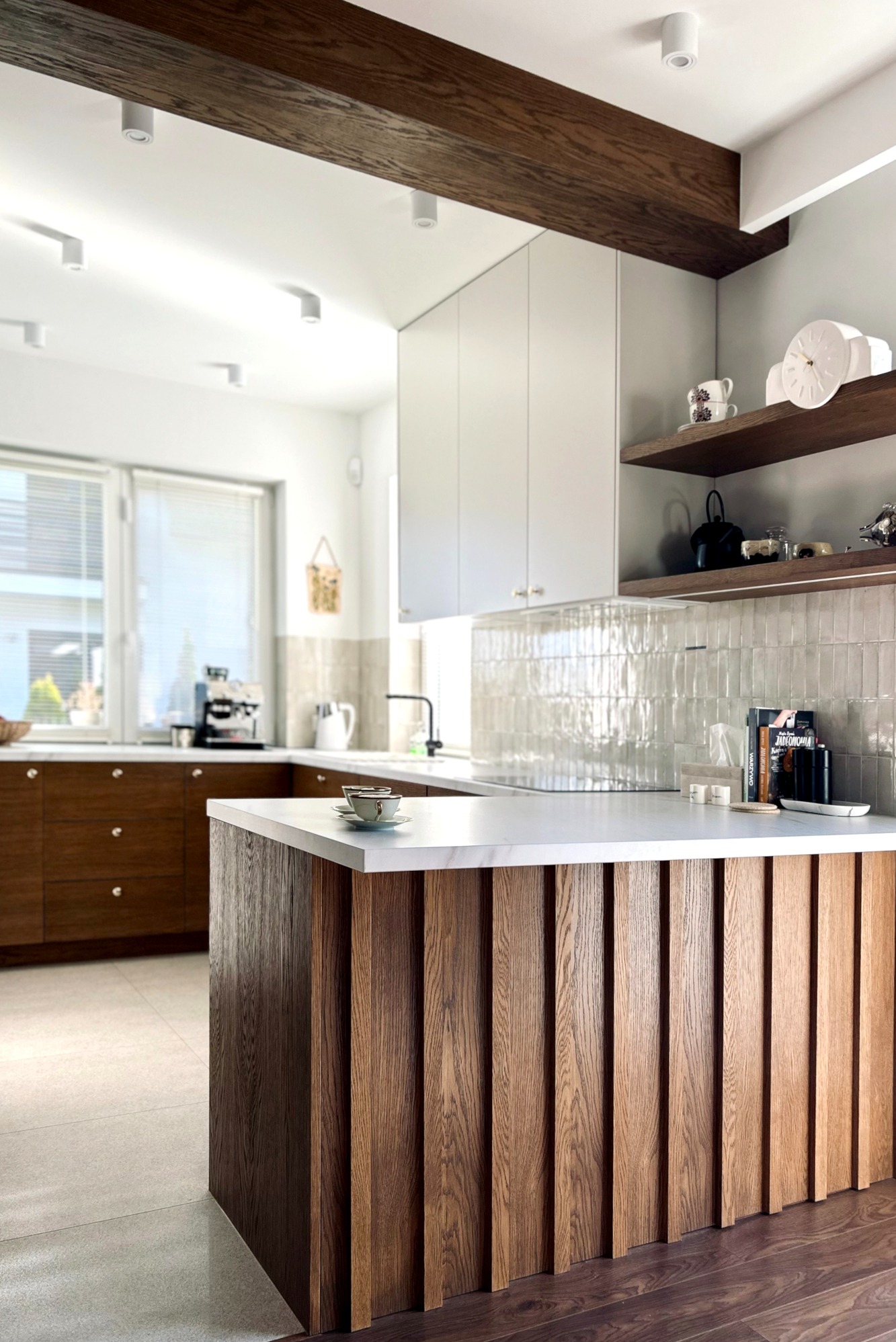 Nowoczesna kuchnia z drewnianą wyspą i białymi szafkami, oświetlona punktowo, z dekoracyjnymi półkami i widokiem na zewnątrz przez okno.