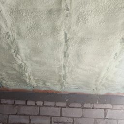 TYNK - BET JUSTYNA SZCZESNA - Poddasze po ociepleniu pianką PUR, widoczne mocowania i struktura piany. Mur z cegieł u dołu kadru. Detal izolacji natryskowej wewnątrz budynku.