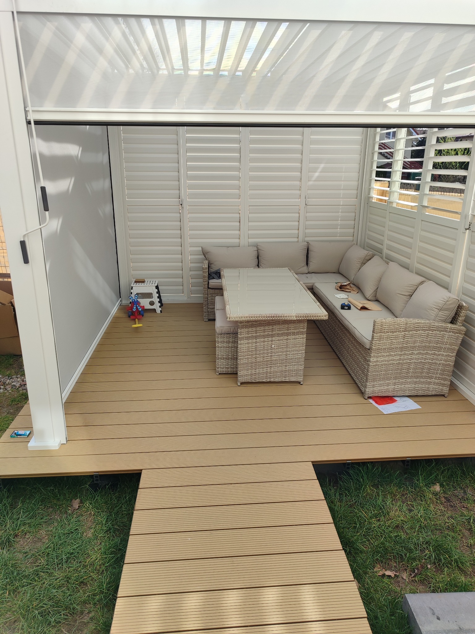 Nowoczesna pergola z meblami ogrodowymi z rattanu i drewnianym podestem. Widoczne detale: zabawka i schodki. Oświetlenie naturalne, jasne kolory, staranne wykończenie.