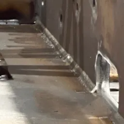 Wnętrze konstrukcji stalowej z widocznymi spawami i otworami. Metal surowy, bez wykończenia, z detalami spoin i śladami po obróbce. Ukazane z bliska, z perspektywą od dołu.