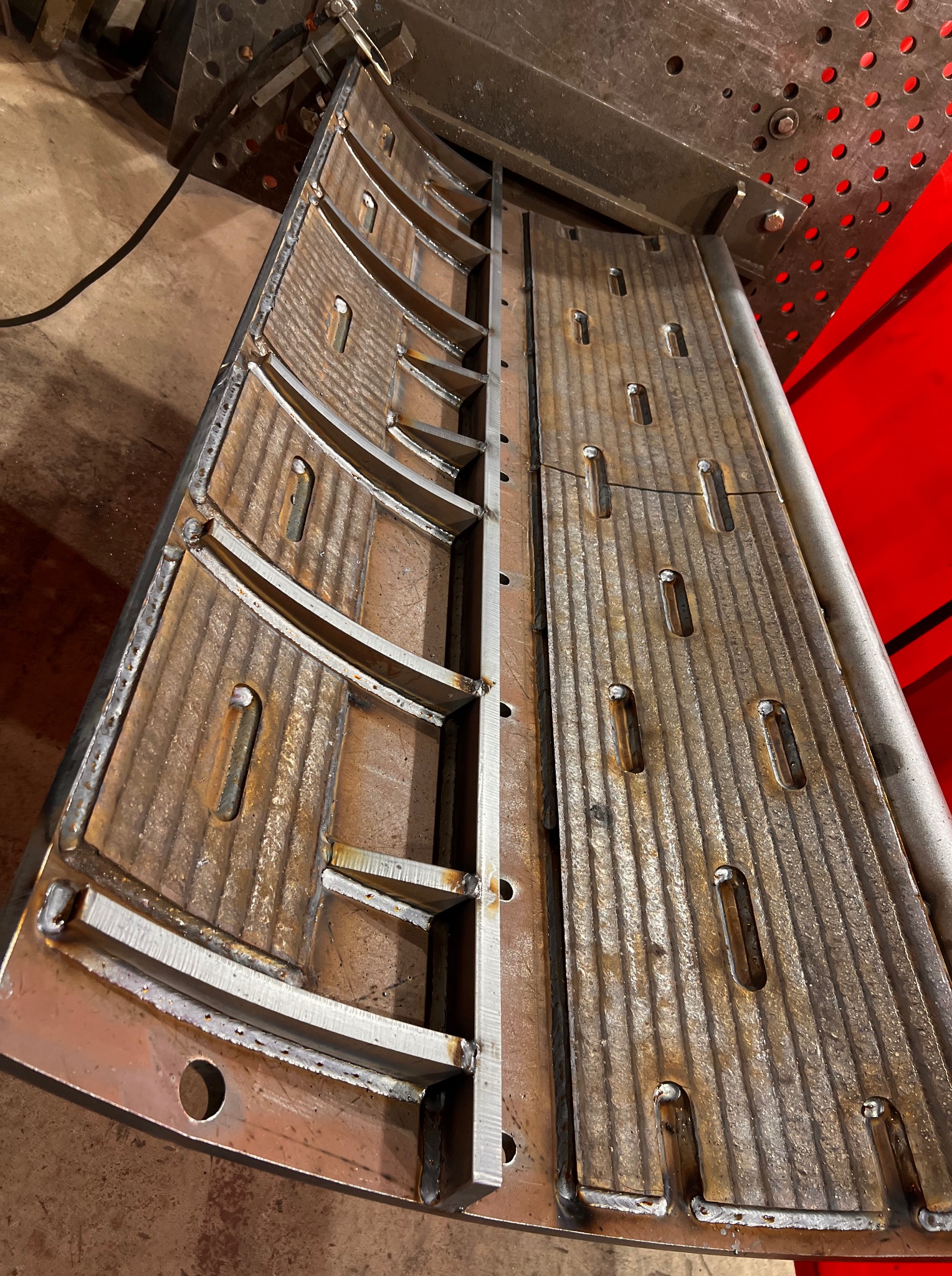 Spawana konstrukcja metalowa z widocznymi śladami spawania, licznymi otworami i żebrowaniem. Element konstrukcyjny na stole spawalniczym, surowy metal, widoczne spawy.