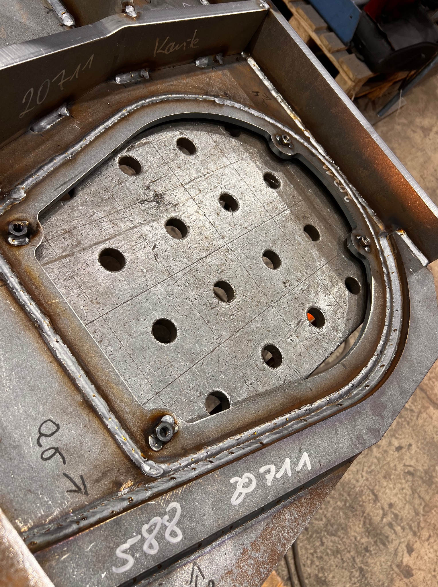 Metalowa konstrukcja z otworami, spawana, z widocznymi śladami obróbki i oznaczeniami. Detal konstrukcyjny w trakcie produkcji, surowy stan.