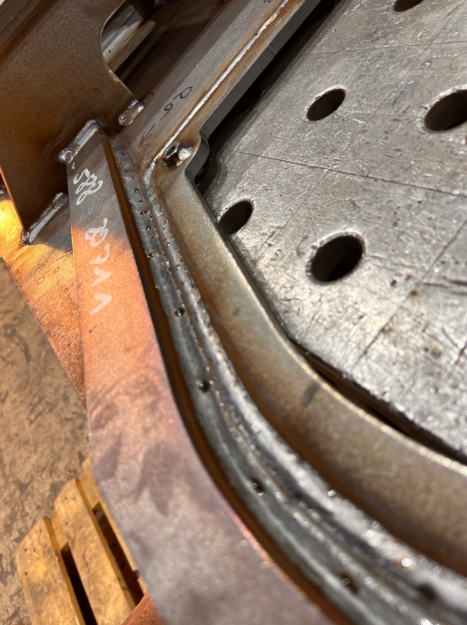 Detale spawania metalowej konstrukcji na stole spawalniczym z otworami, widoczne ślady spawania i napisy na metalu, fragment palety drewnianej w tle.