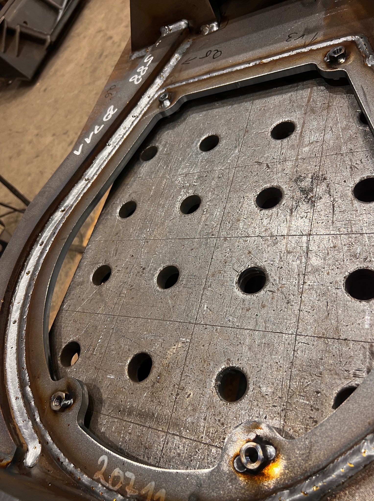 Metalowa konstrukcja z otworami, spawana, z widocznymi śladami obróbki i oznaczeniami. Detal warsztatowy z bliska, surowy wygląd.