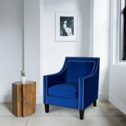Niebieski fotel tapicerowany aksamitem, zdobiony srebrnymi nitami, stoi w minimalistycznym wnętrzu obok drewnianego stolika z kaktusem. Nad nim wisi grafika w białej ramie.