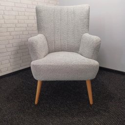 SWARZĘDZKIE PRZEDSIĘBIORSTWO STOLARSKO - TAPICERSKIE RYSZARD TYLKOWSKI - Biały fotel tapicerowany o fakturze baranka na drewnianych nóżkach, na tle ściany z cegły i szarej ściany, na ciemnej wykładzinie dywanowej.