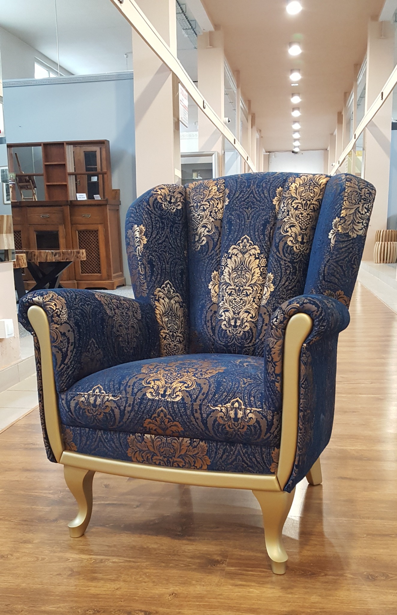 Elegancki fotel uszak w granatowo-złoty wzór damaskowy, z jasnymi nóżkami, na tle wnętrza z drewnianymi meblami i oświetleniem sufitowym.