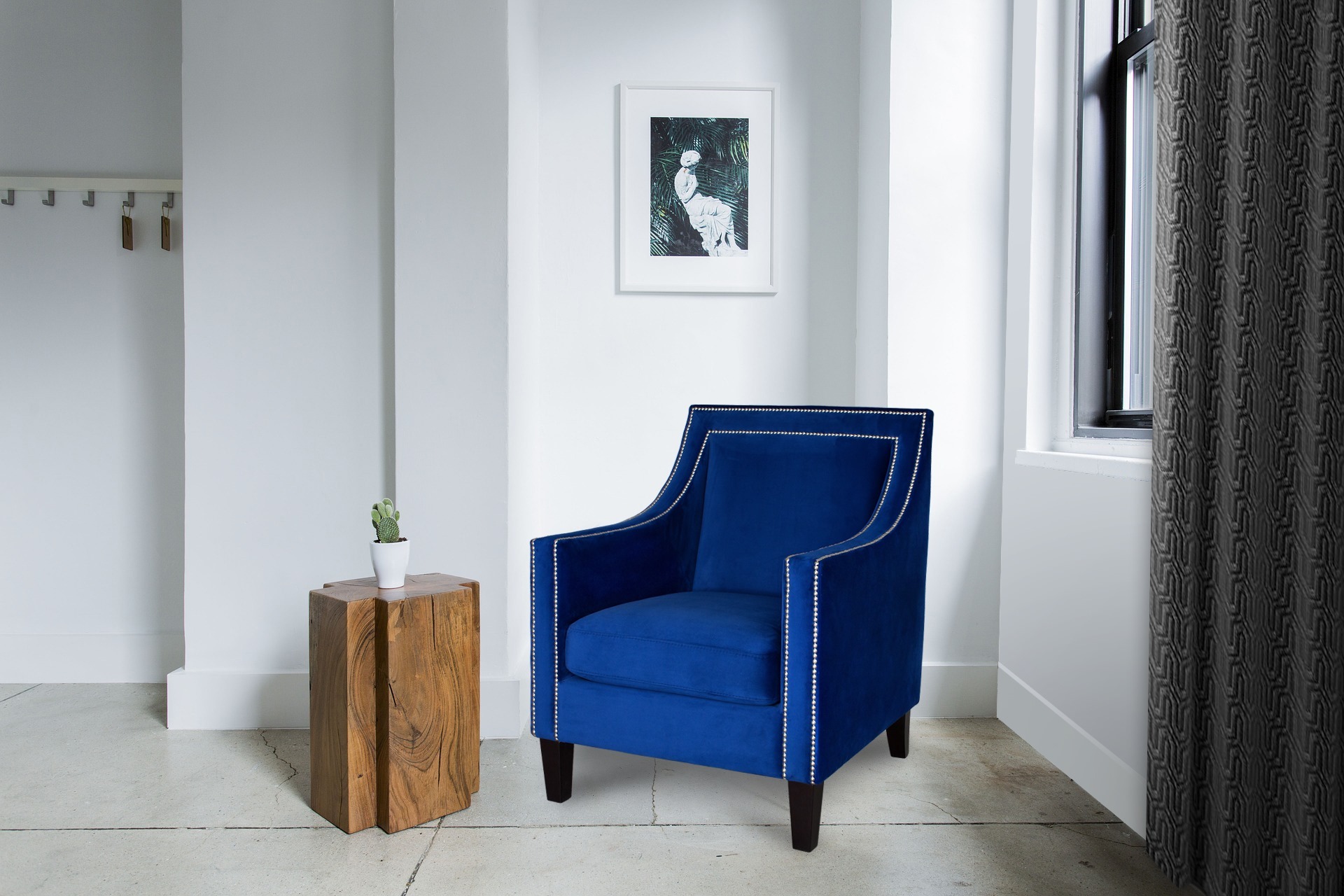 Niebieski fotel tapicerowany aksamitem, zdobiony srebrnymi nitami, stoi w minimalistycznym wnętrzu obok drewnianego stolika z kaktusem. Nad nim wisi grafika w białej ramie.