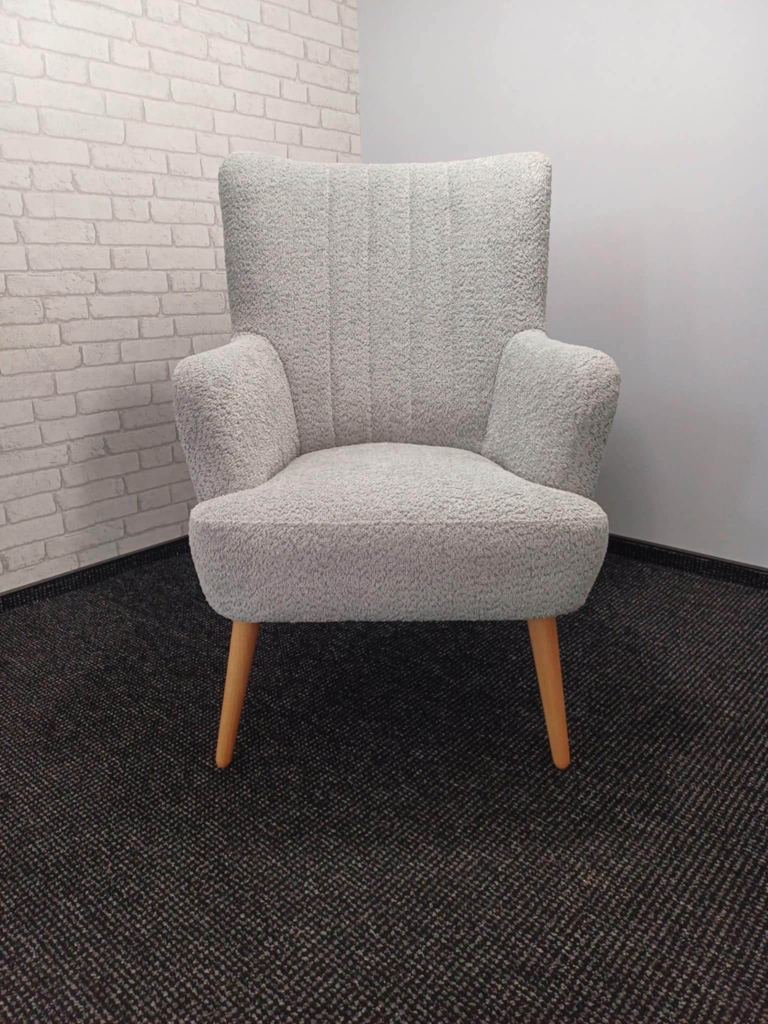 Biały fotel tapicerowany o fakturze baranka na drewnianych nóżkach, na tle ściany z cegły i szarej ściany, na ciemnej wykładzinie dywanowej.