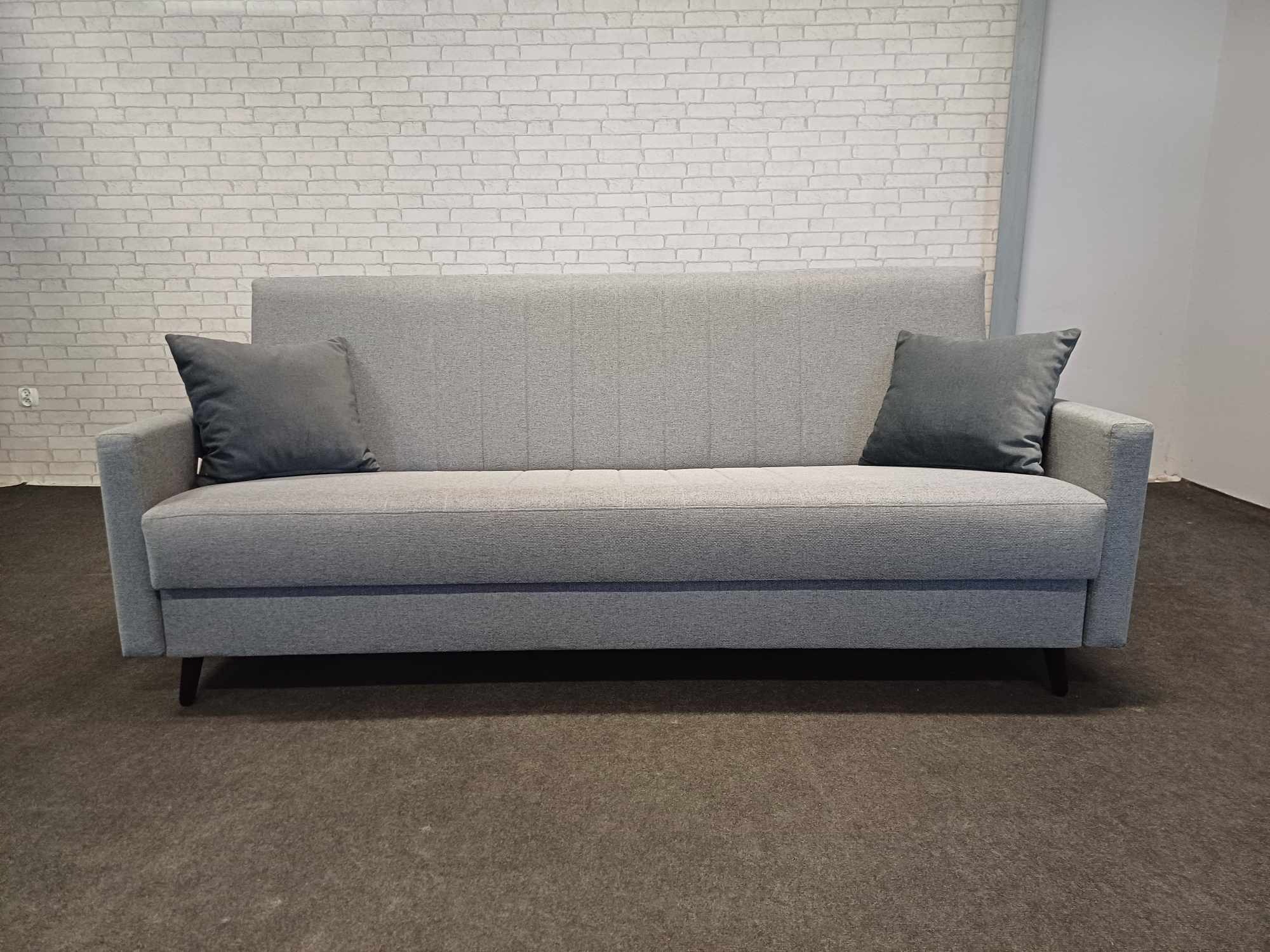Szary, tapicerowany rozkładany narożnik z dwoma poduszkami na tle ściany z białej cegły. Mebel na ciemnych, niskich nóżkach, ustawiony na szarym dywanie.