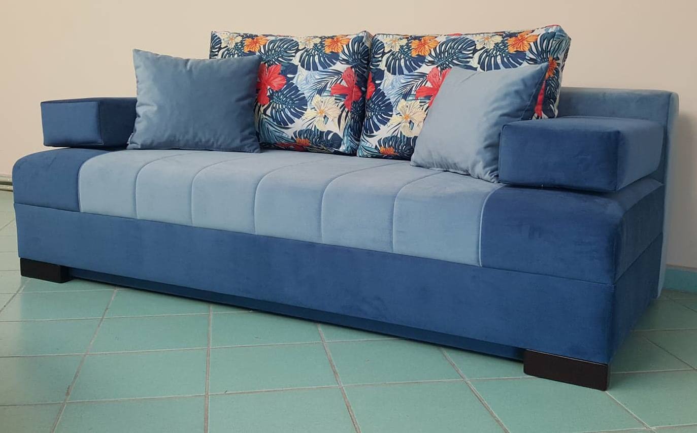 Niebieska sofa z poduszkami w kwiaty, tapicerowana aksamitnym materiałem. Widoczne pikowania na siedzisku i ciemne nóżki. Ujęcie z niskiego kąta.