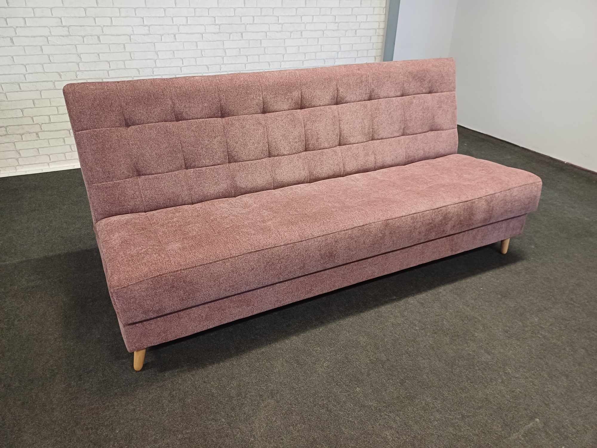 Tapicerowana sofa w odcieniach różu z pikowanym oparciem, na niskich nóżkach. Tło stanowi biała ściana z cegły i szara podłoga.