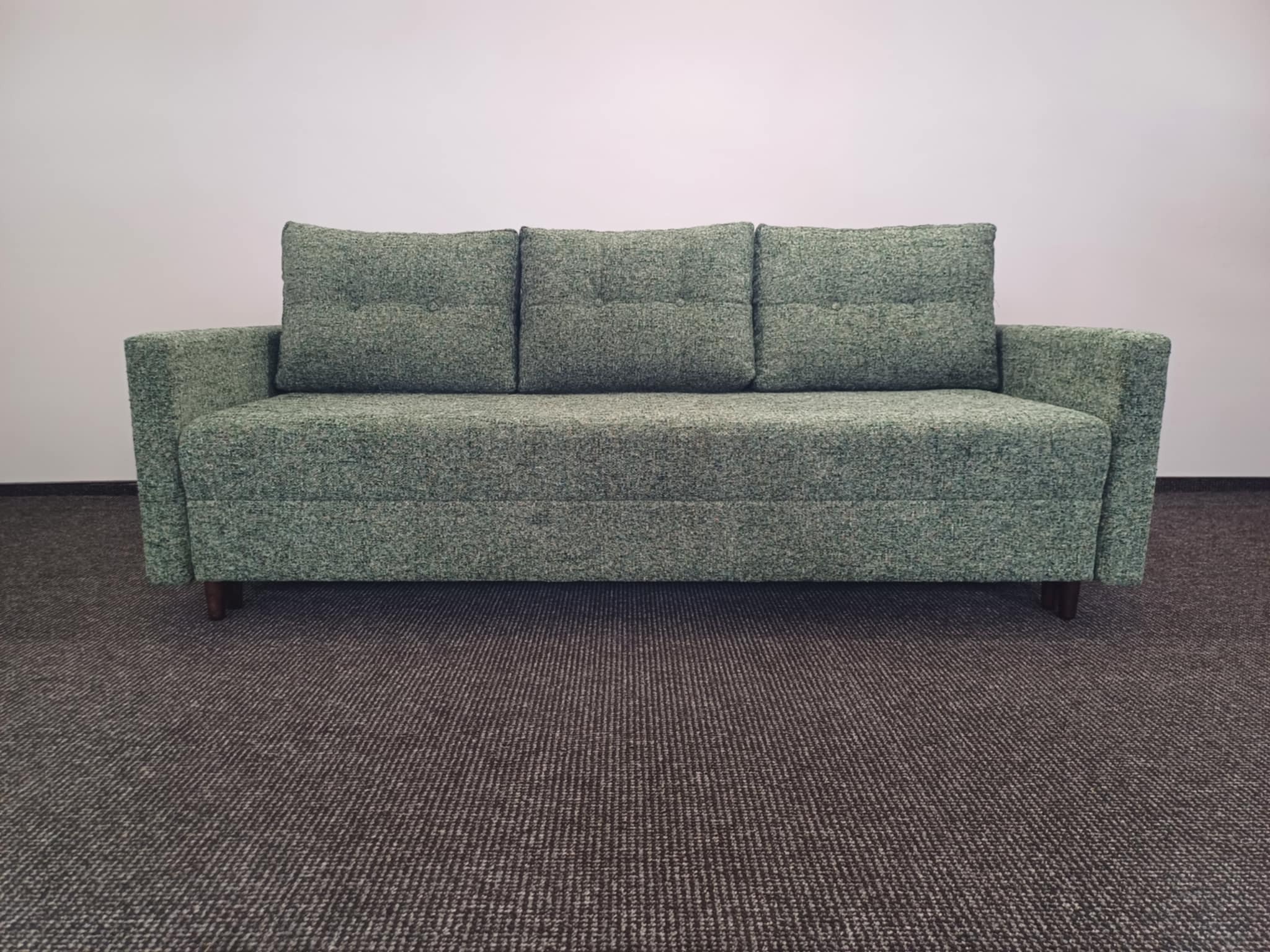 Zielona sofa z trzema poduszkami na siedzisku, tapicerowana tkaniną o wyraźnej fakturze. Widoczne drewniane nóżki i dywan w tle. Prosty, nowoczesny design.