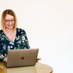 Uśmiechnięta blondynka w okularach, w kwiecistej bluzce, pracuje na laptopie przy okrągłym, złotym stole na tle jasnej ściany. Spokojna atmosfera pracy.