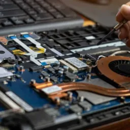 Otwarty laptop z widoczną płytą główną, gdzie dłoń z precyzyjnym śrubokrętem dokonuje naprawy układu chłodzenia, wentylatora.