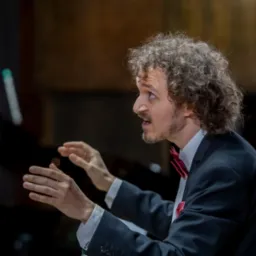 Dyrygent z kręconymi włosami w trakcie prowadzenia orkiestry, ubrany w czarny garnitur i czerwoną muchę, z ekspresją na twarzy i uniesionymi rękami.