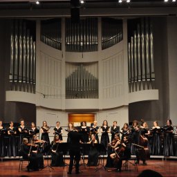 Kasjan Drogosz - Arteterapia, Muzyka i Rozwój - Chór i orkiestra smyczkowa w czarnych strojach występują na scenie z organami w tle, dyrygowani przez mężczyznę w garniturze. Koncert w sali koncertowej.