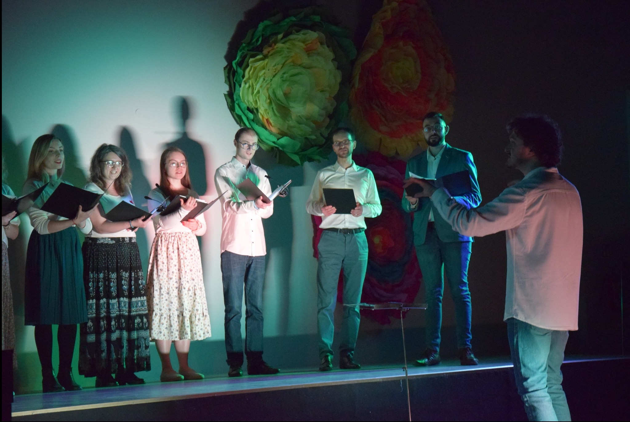 Chór mieszany na scenie, trzymający nuty, oświetlony zielonym światłem, z dekoracjami kwiatowymi w tle i dyrygentem po prawej stronie.