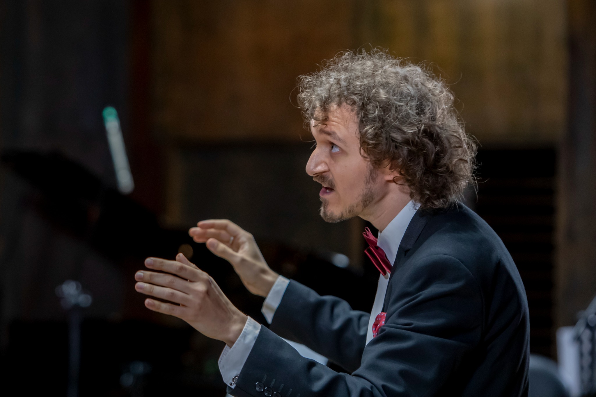 Dyrygent z kręconymi włosami w trakcie prowadzenia orkiestry, ubrany w czarny garnitur i czerwoną muchę, z ekspresją na twarzy i uniesionymi rękami.