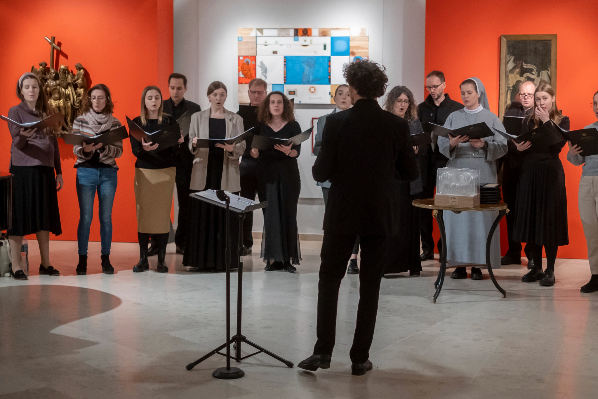 Chór mieszany w trakcie występu w galerii sztuki, dyrygent stoi przodem do chóru, w tle pomarańczowa ściana i obrazy.