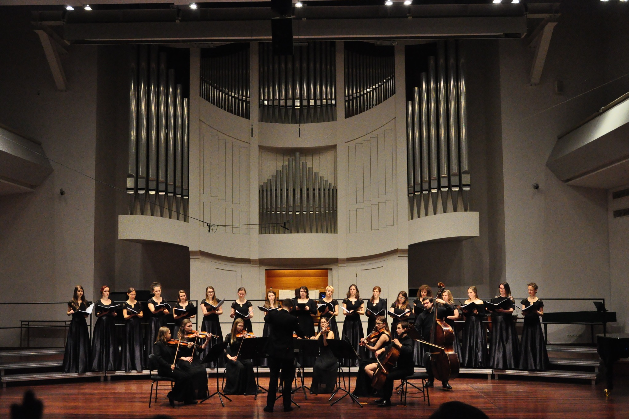 Chór i orkiestra smyczkowa w czarnych strojach występują na scenie z organami w tle, dyrygowani przez mężczyznę w garniturze. Koncert w sali koncertowej.