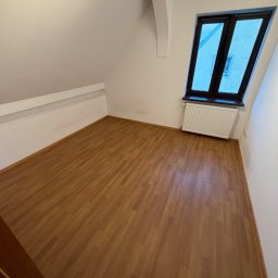 Patrex Patryk Bejynka - Puste poddasze z drewnianą podłogą, białymi ścianami i oknem. Widoczny grzejnik pod oknem. Perspektywa z narożnika pokoju, drzwi częściowo widoczne po lewej.
