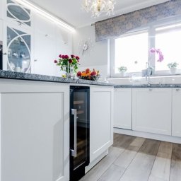 Patrex Patryk Bejynka - Biała kuchnia z wyspą i wbudowaną chłodziarką na wino. Na blacie granitowym owoce i kwiaty. Okno z roletą rzymską. Parkiet w odcieniach szarości.