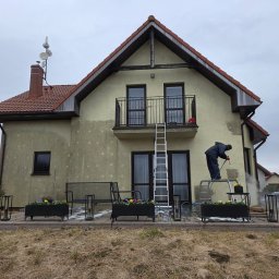 Patrex Patryk Bejynka - Czyszczenie elewacji domu jednorodzinnego myjką ciśnieniową. Widoczny fragment wyczyszczonej ściany, drabina i pracownik w kombinezonie ochronnym.