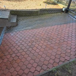Patrex Patryk Bejynka - Częściowo umyta, czerwona kostka brukowa na podjeździe, z widocznym odpływem liniowym i fragmentem ogrodzenia. Górna część kostki jest jeszcze brudna.