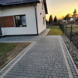 Patrex Patryk Bejynka - Ułożona kostka brukowa w odcieniach szarości prowadząca do nowoczesnego domu z białymi ścianami i drewnianymi akcentami, widok na ogród i zachodzące słońce.