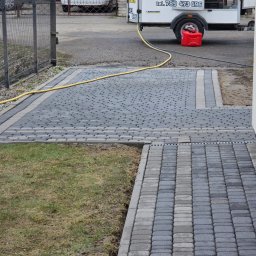 Patrex Patryk Bejynka - Wyczyszczona kostka brukowa w odcieniach szarości, z widocznym w tle sprzętem do mycia ciśnieniowego i żółtym wężem. Część kostki poza kadrem.