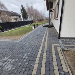 Patrex Patryk Bejynka - Ułożona kostka brukowa w dwóch odcieniach szarości, z jasnymi pasami akcentującymi ścieżkę. Widoczny fragment domu z białymi ścianami i czarnymi elementami wykończenia.