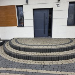 Patrex Patryk Bejynka - Ułożona kostka brukowa przed domem z numerem 11, tworząca półokrągłe schody wejściowe. Szara, nowoczesna stolarka okienna i drzwi. Elementy drewniane na elewacji.