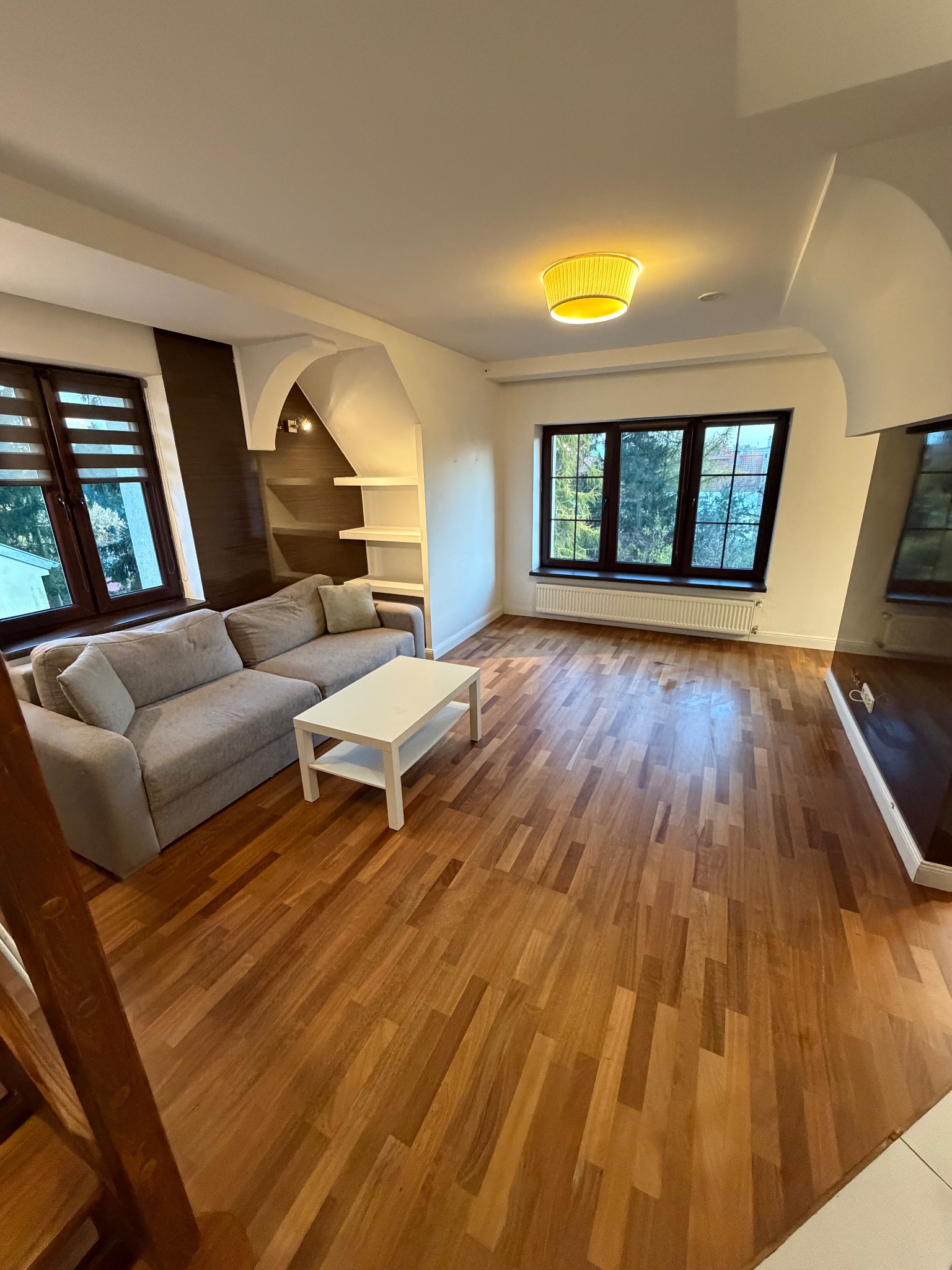 Jasne wnętrze po remoncie: parkiet, szara sofa, biały stolik kawowy, okna z roletami, lampa z abażurem. Widok na czyste, odświeżone pomieszczenie.