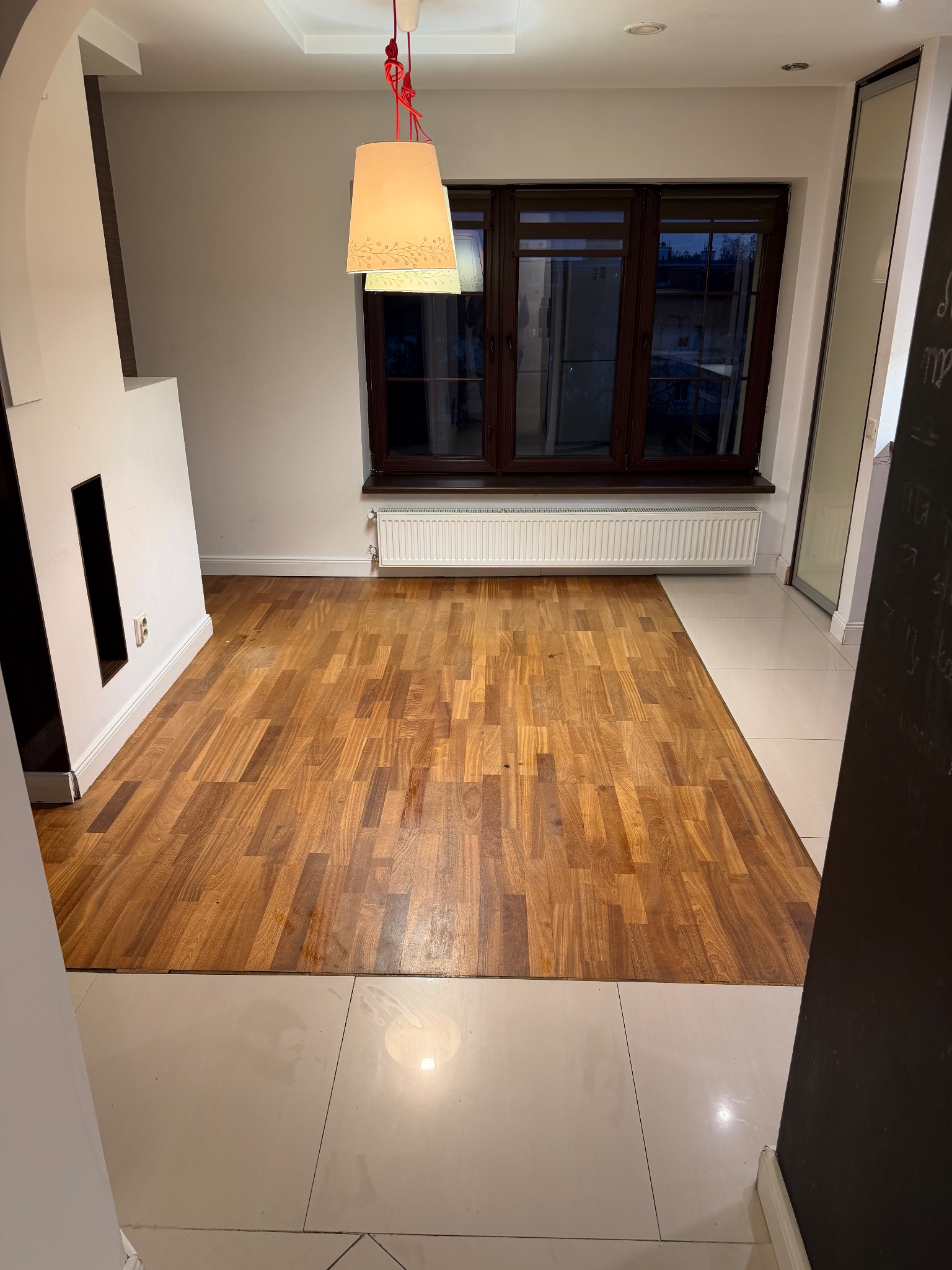 Wnętrze pokoju po sprzątaniu: parkiet z drewna ułożony w jodełkę, białe płytki podłogowe, okno z brązową ramą, biały grzejnik i nowoczesny kominek.