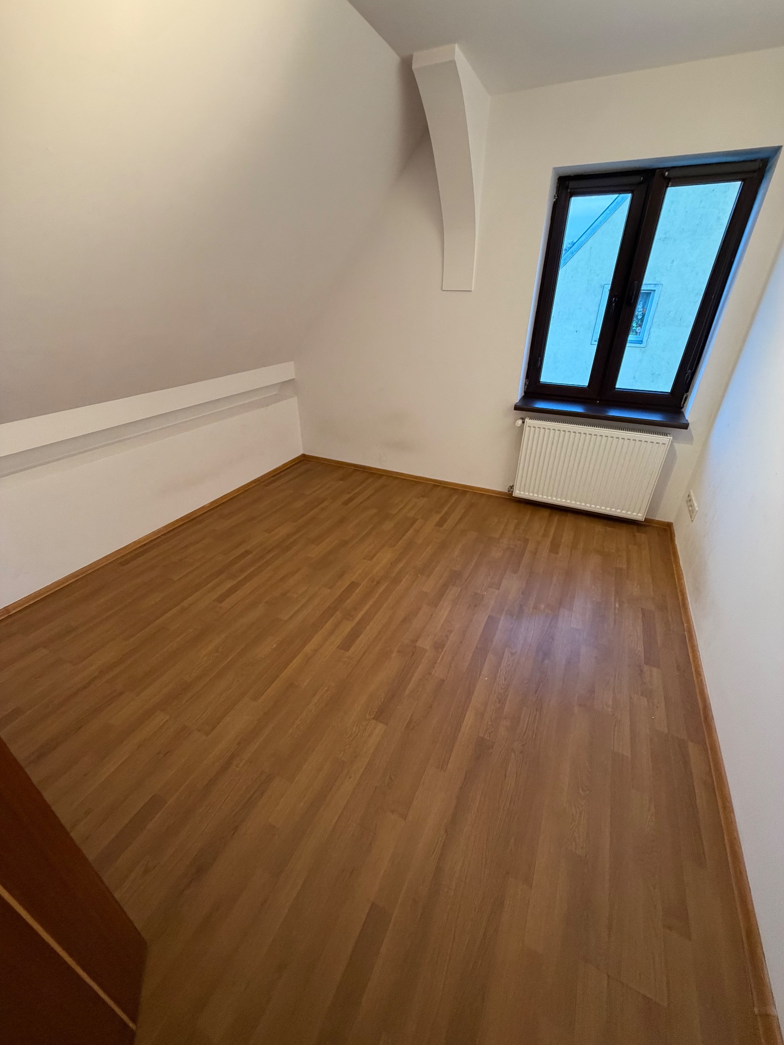 Puste poddasze z drewnianą podłogą, białymi ścianami i oknem. Widoczny grzejnik pod oknem. Perspektywa z narożnika pokoju, drzwi częściowo widoczne po lewej.